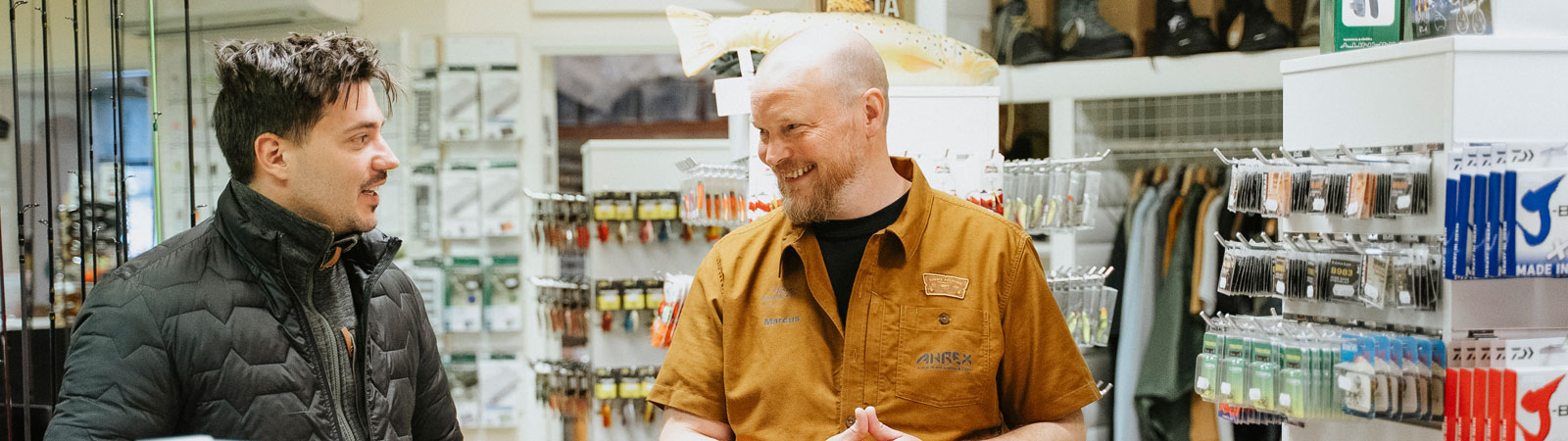 Två män pratar i en fiskebutik med regnbåge på väggen och flugor i hyllor.