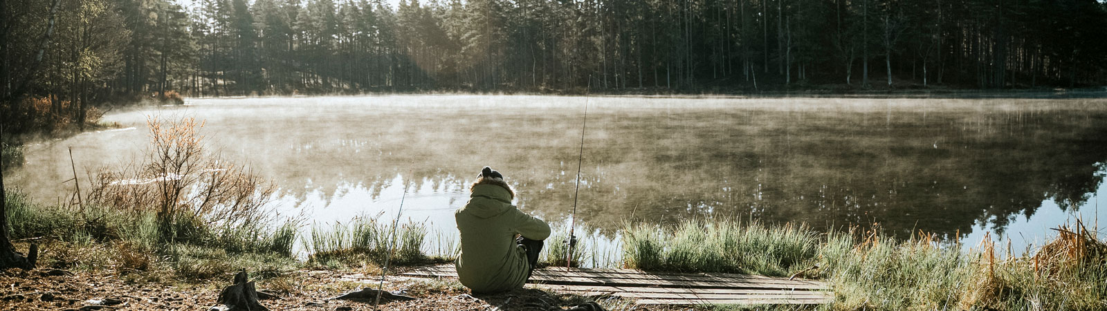 En person sitter vid en dimmig sjö och fiskar med två spö i morgonljus.