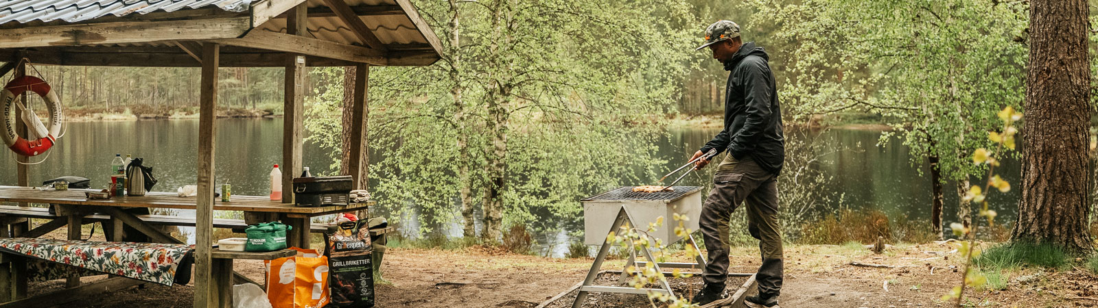 En man grillar fisk vid en skogsbrygga med en trähylla och en sjö i bakgrunden.