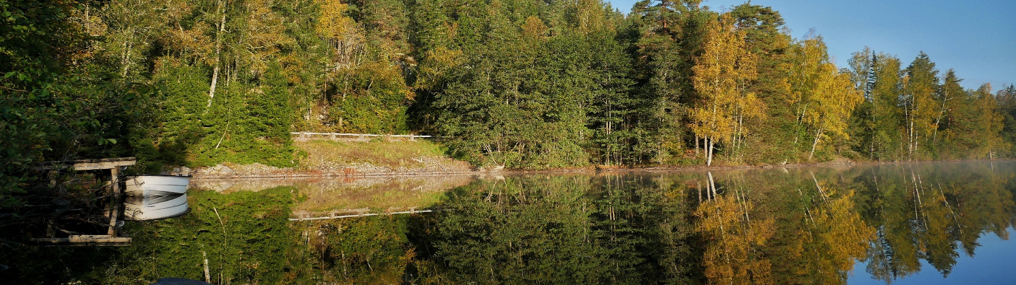 En stilla sjö med spegelblank yta, omgiven av höstfärgad skog och en vit båt vid kajen.