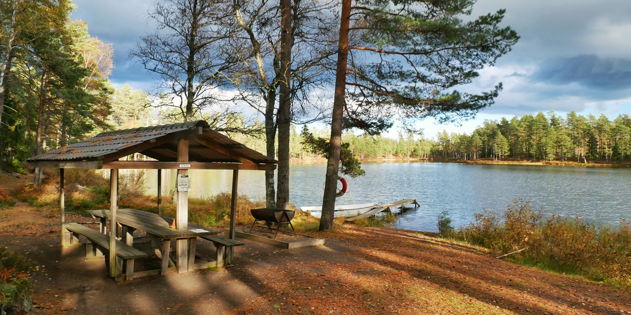 En träsköld med bänkar vid en stilla sjö omgiven av höstfärgade träd och en liten brygga.