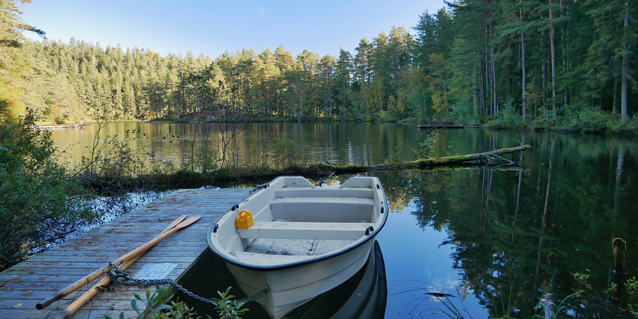 En vit båt ligger vid en träkaj vid ett stilla sjö med skog i bakgrunden.