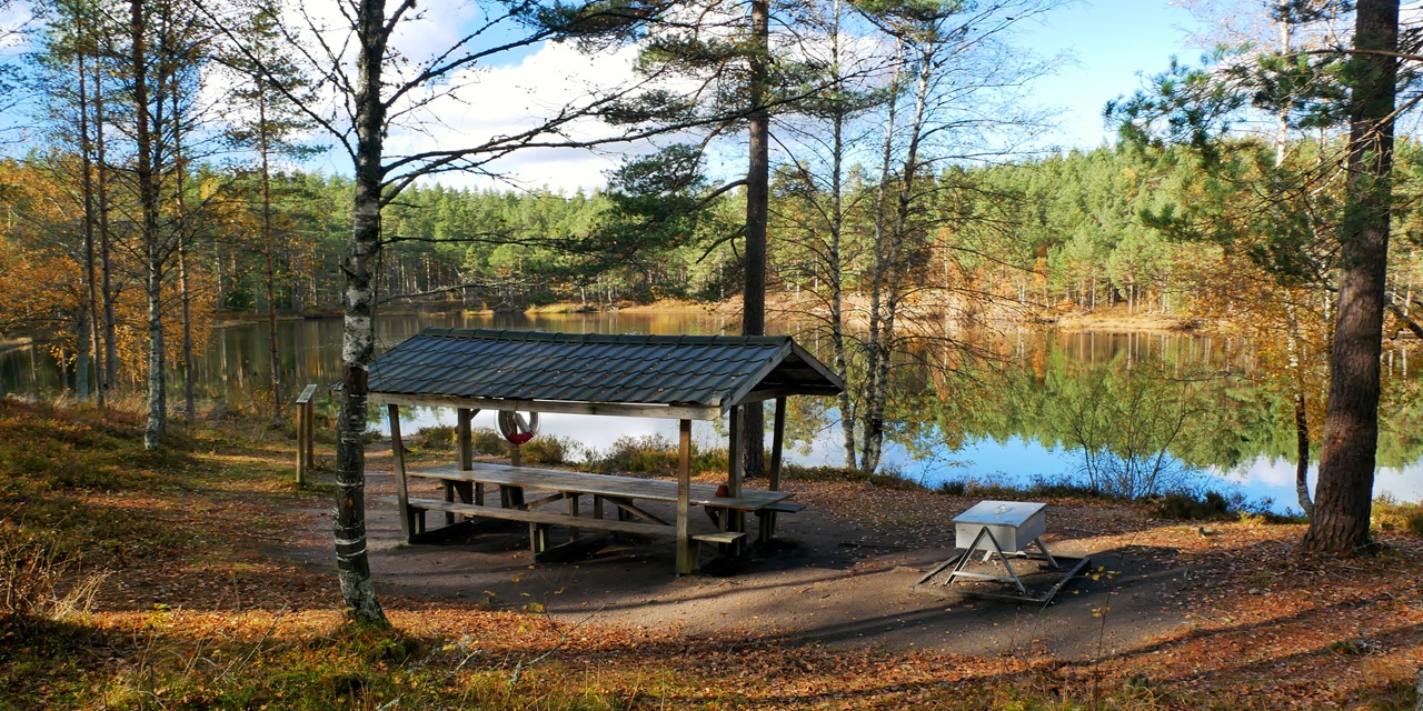 En träpaviljong vid en stilla sjö omgiven av höstfärgade träd och reflekterande vatten.