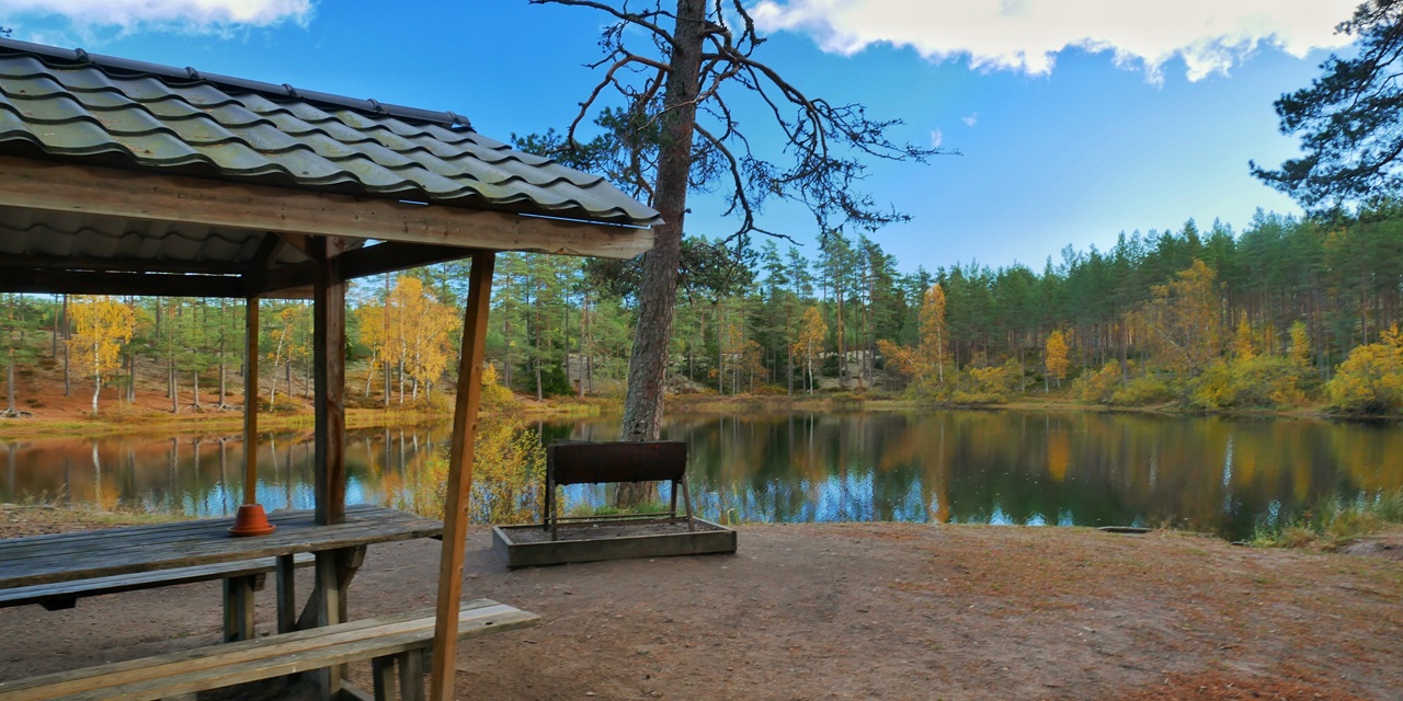 En trähylla vid en stilla sjö med höstfärgade träd och klar blå himmel.