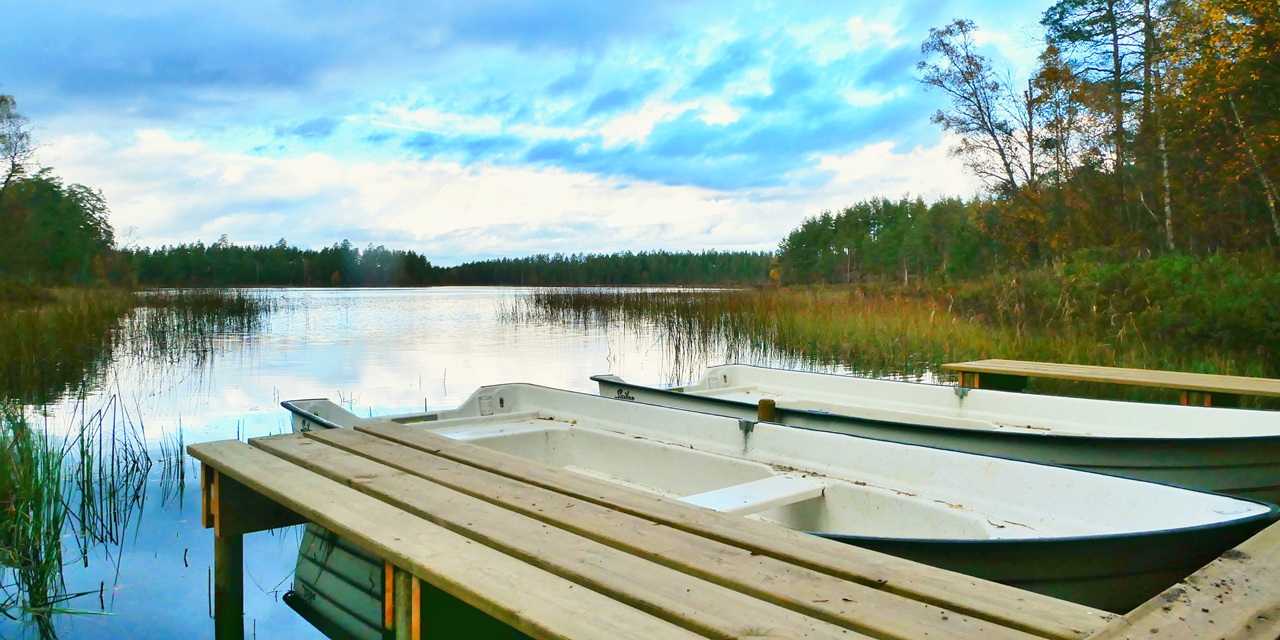 Två vita fiskebåtar vid en träkaj på ett stilla sjölandskap med skog och moln.