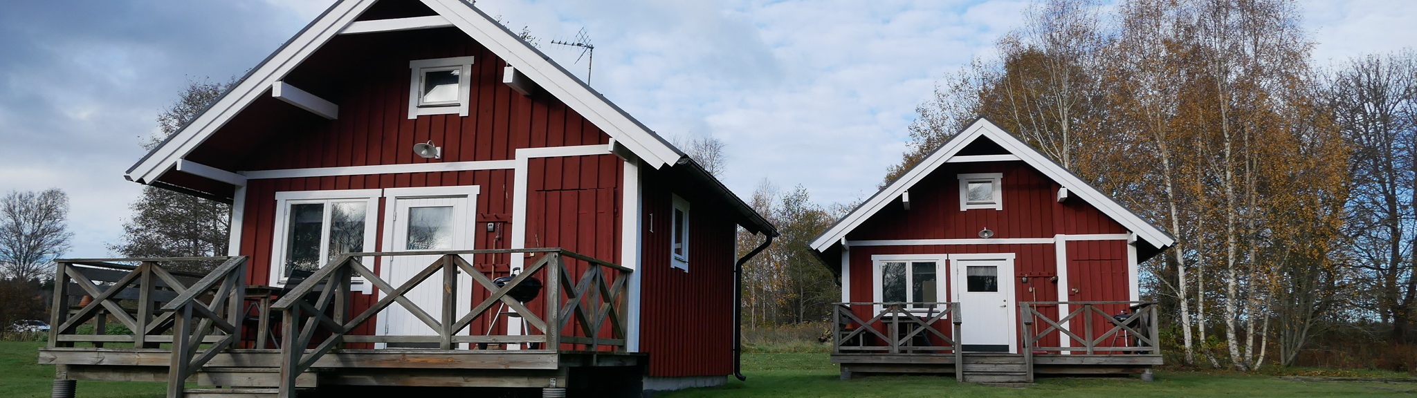 Två röda trästugor med vita detaljer står på gräs, omgivna av höstträd.