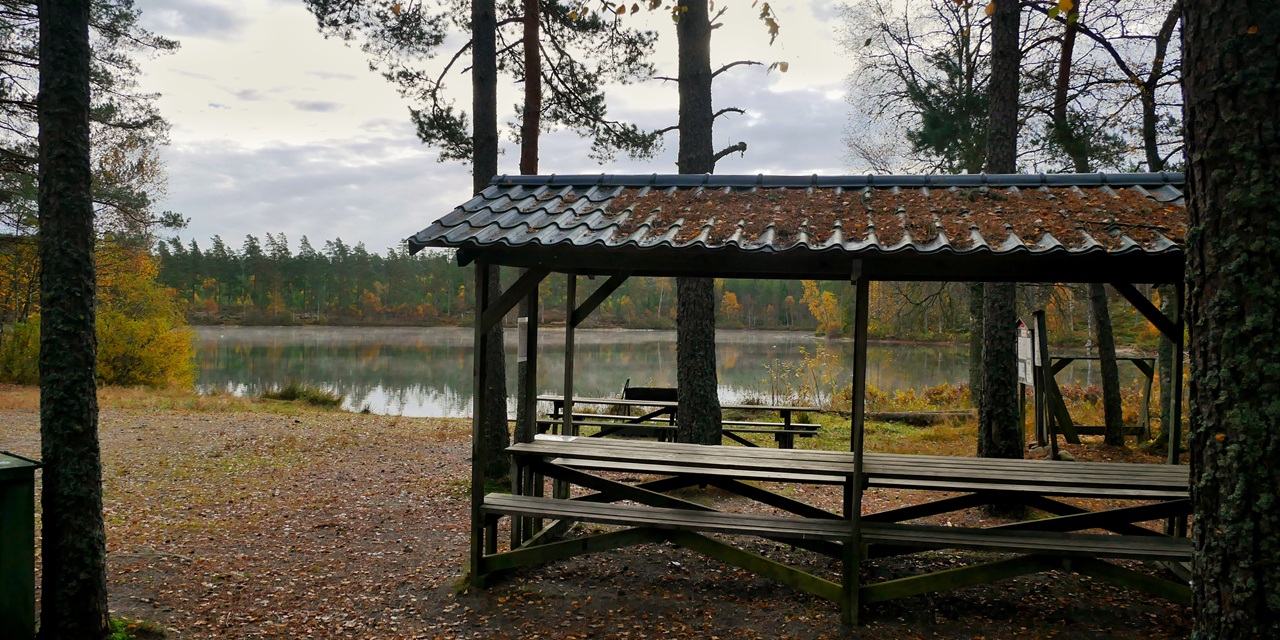 En trägångstuga med bänkar vid en stilla sjö omgiven av höstfärgade träd.