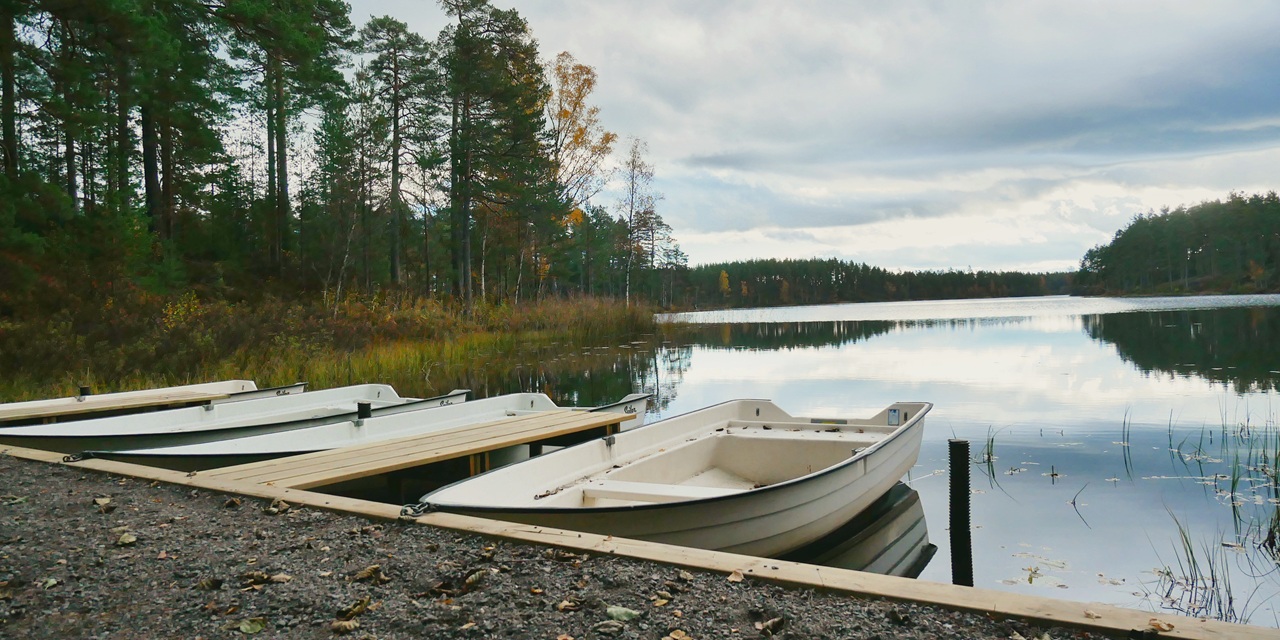 Vit båt vid kaj på en stilla sjö med skog i bakgrunden under molnig himmel.