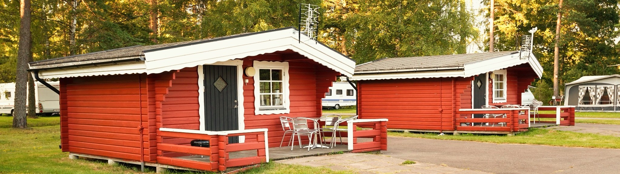Röda trästugor med vita detaljer i en skogsmiljö, med campingvagnar i bakgrunden.